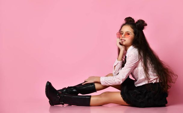 Teenage child with long hair, in sunglasses, white blouse, black skirt and boots. Posing sitting on pink background. Close up - Fotografie, Obrázek