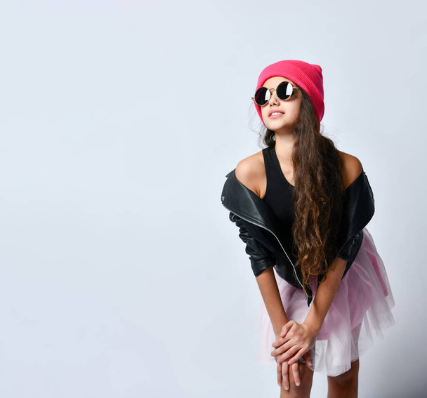 Teenager in black leather jacket, skirt, pink hat and sunglasses. She smiling while posing standing isolated on white. Close up - Фото, зображення