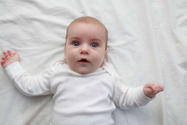 A baba mosolyog és egy fehér pulóverben fekszik egy fehér lepedőn, kinyújtott karokkal. Baba gyönyörű kék szemekkel. - Fotó, kép