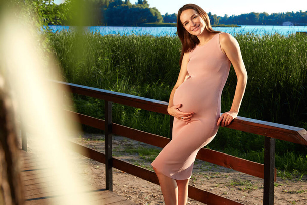 Beautiful young pregnant woman in anticipation of a newborn at 9 month of pregnancy walking in the park tender summer weather green trees dressed in a tight-fitting pink dress holding onto the belly. - Photo, image