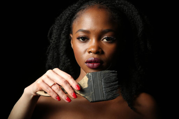 Portré egy fiatal, gyönyörű afrikai nőről, sűrű fekete göndör hajjal, fekete háttérrel. Mellette egy kéz egy fekete festékkel festett ecsetet tart.. - Fotó, kép