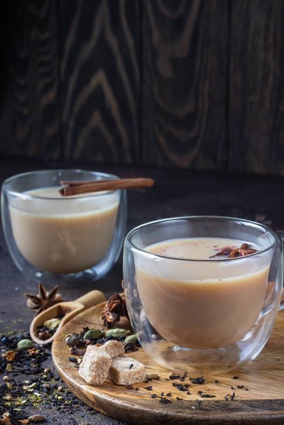 Traditional masala chai in glass cup with ingredients (cinnamon, anise, cardamom, nutmeg, pepper and brown sugar) on a dark stone background.  - Foto, imagen