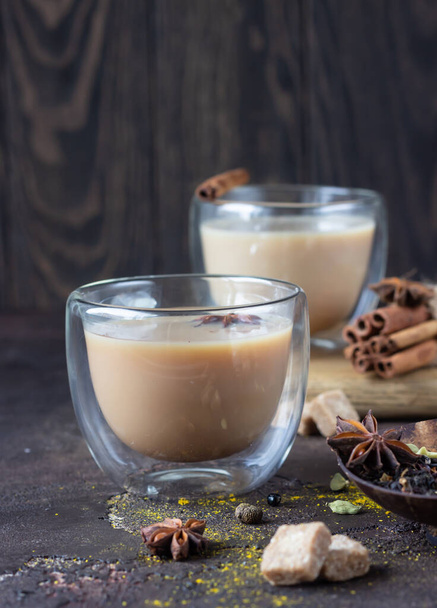 Traditional masala chai in glass cup with ingredients (cinnamon, anise, cardamom, nutmeg, pepper and brown sugar) on a dark stone background.  - Valokuva, kuva