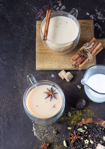 Traditional masala chai in glass cup with ingredients (cinnamon, anise, cardamom, nutmeg, pepper and brown sugar) on a dark stone background.  - Photo, image
