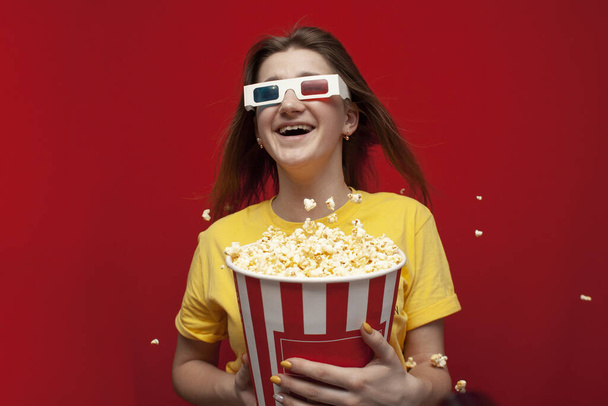 funny young girl shocked in 3D glasses watching a movie and eating popcorn on a red colored background, she smiles and popcorn flies, copy space - Photo, Image