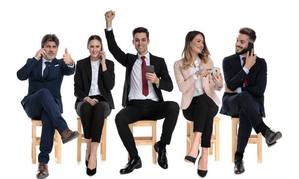 Ομάδα 5 επιχειρηματιών που γιορτάζουν και κρατούν τα τηλέφωνά τους ενώ κάθονται σε καρέκλες σε λευκό φόντο στούντιο - Φωτογραφία, εικόνα