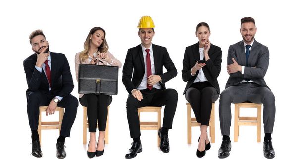 Team of 5 businessmen thinking, wearing safety helmet and reading from phone waiting for a job interview sitting on chairs on white studio background - Photo, Image