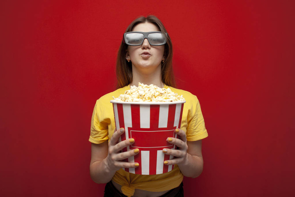 happy young girl the viewer in 3D glasses holds a big box of popcorn and watches a movie on a red color background, copy space - Photo, image