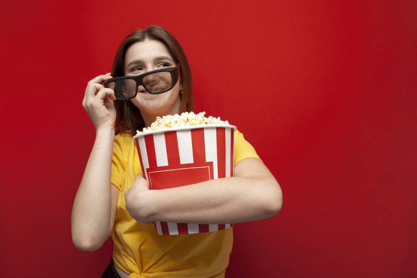 happy hipster girl with popcorn puts on 3D glasses and smiles on a red colored background, copy space - Photo, Image