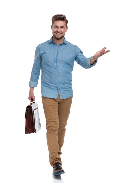 Smiling casual man presenting while holding briefcase and stepping on white studio background - Foto, Imagen
