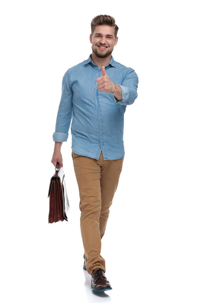 Positive casual man giving thumbs up while holding briefcase and stepping on white studio background - Fotó, kép