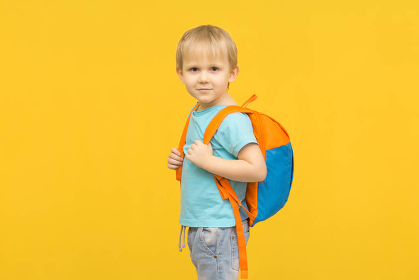 Χαρούμενο παιδί στέκεται με ένα σακίδιο στην πλάτη και κοιτάζει την κάμερα σε ένα φωτεινό κίτρινο φόντο. Ιδέα για ταξιδιωτικά είδη και άνετα βρεφικά ρούχα. - Φωτογραφία, εικόνα
