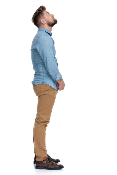 Vista lateral de um homem casual pensativo olhando para cima e se perguntando, de pé no fundo do estúdio branco
 - Foto, Imagem