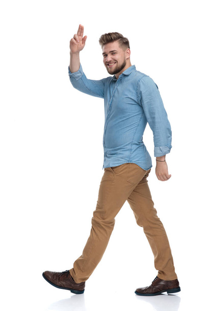 Side view of a cheerful casual man greeting and laughing while stepping on white studio background - 写真・画像