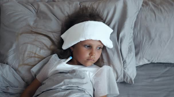Sick girl with a thermometer and compress on forehead lying in bed - Footage, Video