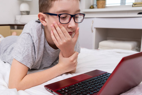 A boy wearing glasses lying in bed and using a laptop, typing the text on a keyboard close up, online education concept. - Φωτογραφία, εικόνα
