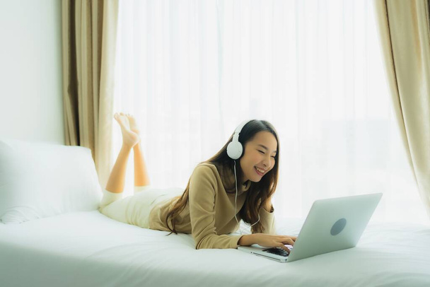 Genç Asyalı kadın, yatak odasının içinde müzik dinlemek için kulaklıklı laptop kullanıyor. - Fotoğraf, Görsel