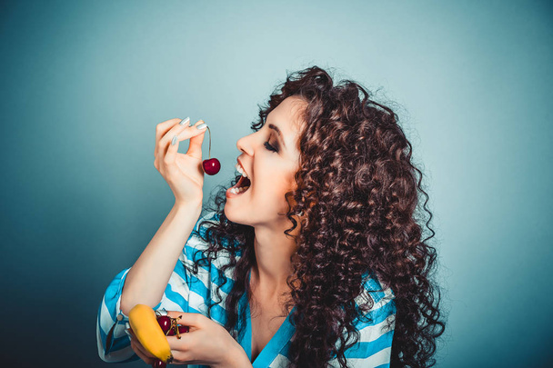 Healthy eating concept. Attractive young beautiful curly woman holding enjoying cherry in open mouth with eyes closed holding banana in other hand happy isolated on blue background wall with vignette  - Foto, imagen