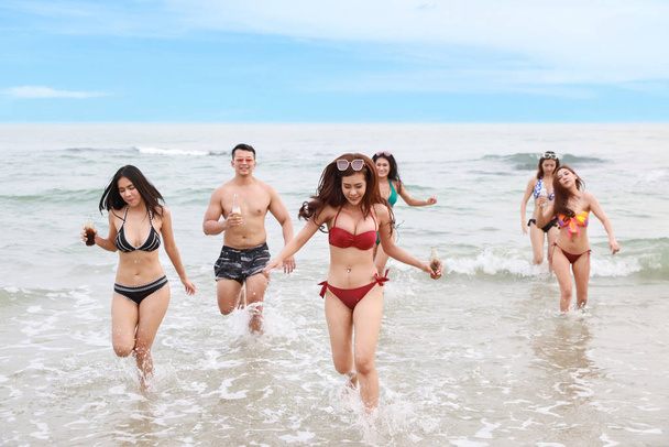 group of happy and sexy asian friends who wearing bikini and swimming suit. they running and holding alcohol and soft drink bottle on sandy beach party. young people having fun and happiness smiling face. - Photo, Image