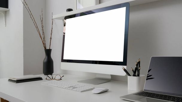Vista ravvicinata di uno spazio di lavoro minimo con computer con schermo bianco, laptop, cancelleria e decorazioni su scrivania bianca con mensola su parete bianca
  - Foto, immagini