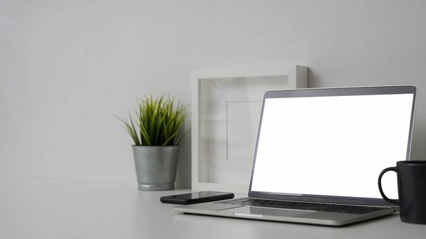 Gehakt schot van comfortabele werkruimte met blanco scherm laptop en decoraties op witte tafel met witte wand achtergrond   - Foto, afbeelding