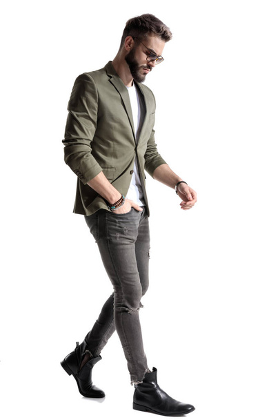 Vue latérale d'un mannequin réfléchi tenant sa main dans sa poche tout en portant une veste verte et des lunettes de soleil, marchant sur fond de studio blanc
 - Photo, image