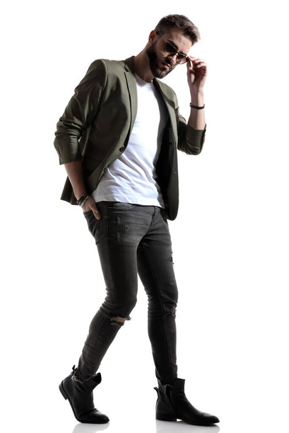 Charmant mode model vaststelling van zijn zonnebril en houdt zijn hand in zijn achterzak, stappen op witte studio achtergrond - Foto, afbeelding