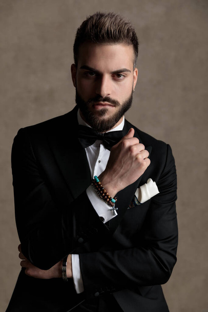 Tough groom biting his lip and holding his fist clenched while wearing tuxedo and sitting on a stool on wallpaper studio background - Zdjęcie, obraz
