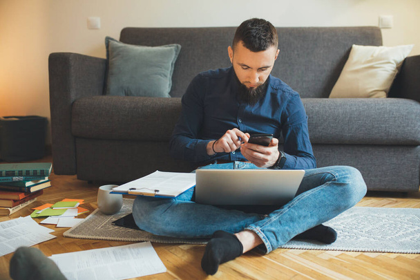 Drukke blanke freelancer draagt een shirt en mooie baard werkt hard zittend op de vloer met een laptop en wat documenten - Foto, afbeelding