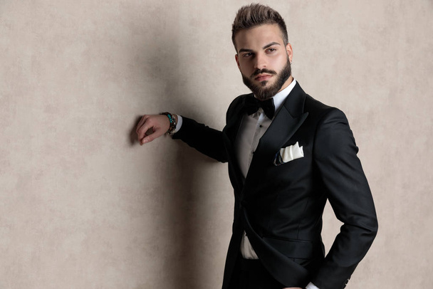 Tough groom looking away with his hand in his pocket while wearing tuxedo and standing on wallpaper studio background - Fotografie, Obrázek