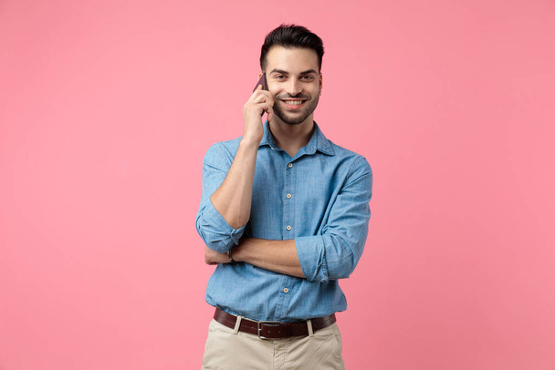 Ευτυχισμένος νεαρός με τζιν πουκάμισο να μιλάει στο τηλέφωνο και να στέκεται σε ροζ φόντο. - Φωτογραφία, εικόνα