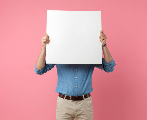 casual guy in denim shirt covering face with empty board, standing on pink background - 写真・画像