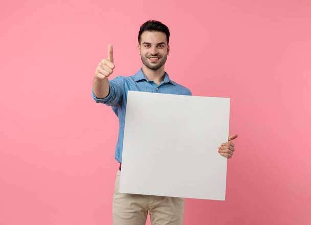 young casual guy making thumbs up sign, holding empty board and smiling, standing on pink background - Photo, Image