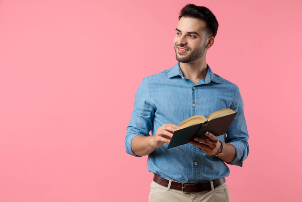 young casual guy holding book and looking to side, standing on pink background - Фото, изображение