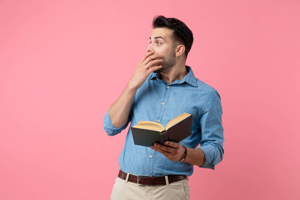 šokovaný ležérní chlap čtení knihy a drží ruku v puse, hledá na straně na růžovém pozadí - Fotografie, Obrázek