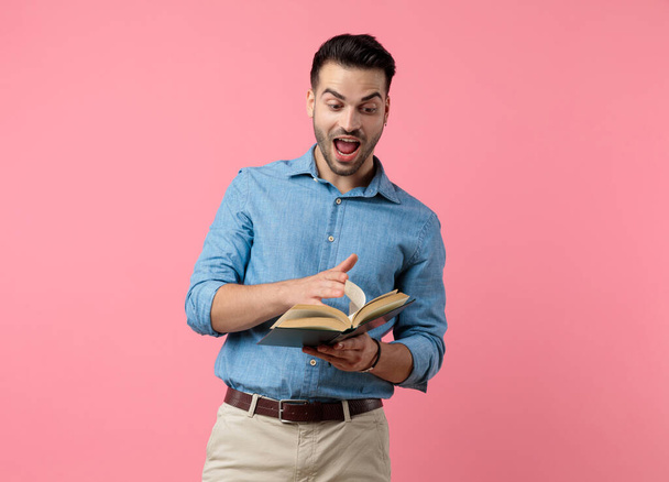 happy young guy laughing, holding book and flipping pages, standing on pink background - Foto, Imagen