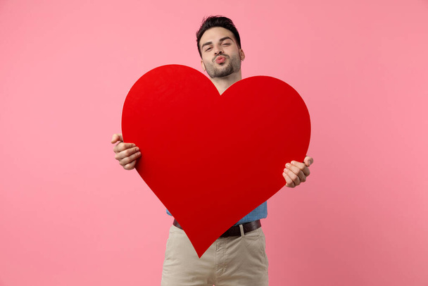 happy young guy holding big red heart sending kiss and standing on pink background - Foto, Imagen