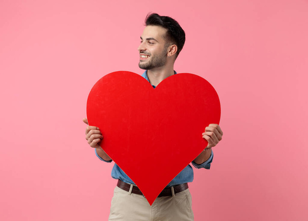 happy young guy smiling and holding big red heart, standing on pink background  - Zdjęcie, obraz