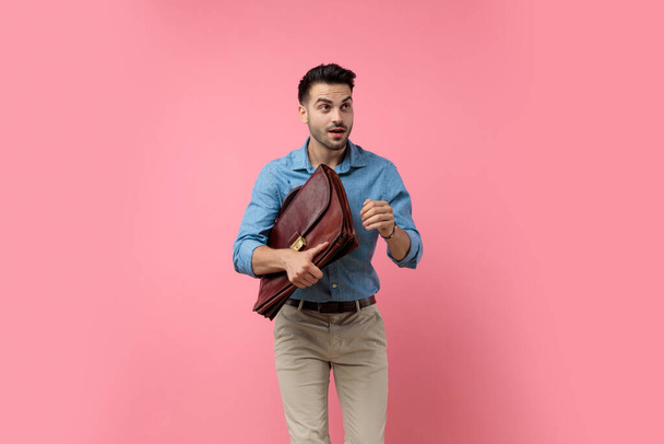 młody casual facet w dżinsowej koszuli patrząc w górę i trzymając walizkę na różowym tle, jest gotowy do skoku - Zdjęcie, obraz