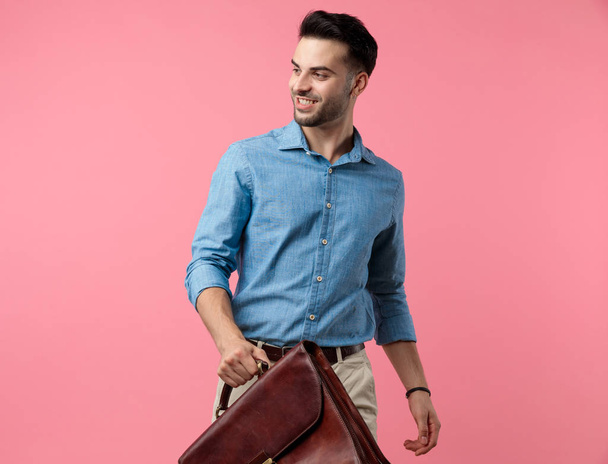 happy young guy in denim shirt smiling and holding suitcase, standing on pink background - Fotó, kép