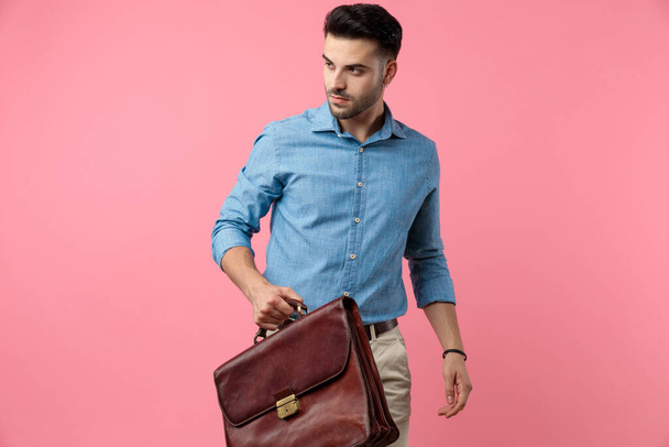 sexy jonge man op zoek naar kant en het houden van koffer, lopen op roze achtergrond - Foto, afbeelding