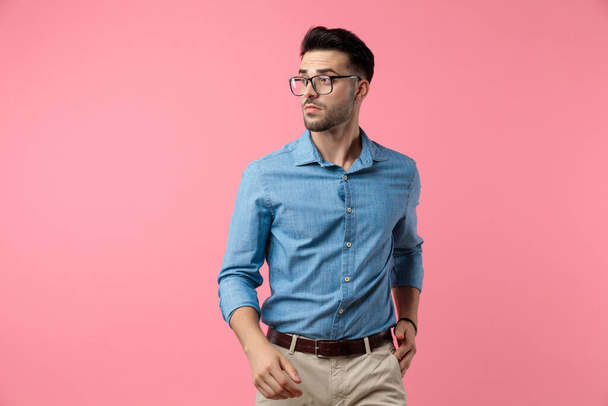 σέξι νεαρός άνδρας σε denim πουκάμισο κοιτάζοντας προς τα πλάγια και να ποζάρουν, περπατώντας σε ροζ φόντο - Φωτογραφία, εικόνα