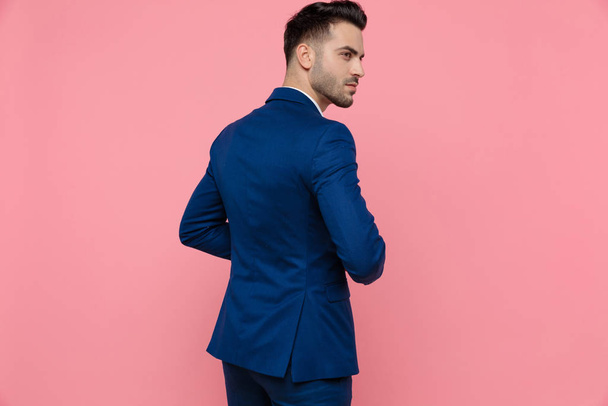 Πίσω όψη ενός γοητευτικού επιχειρηματία που φοράει μπλε κοστούμι και στέκεται σε ροζ φόντο στούντιο - Φωτογραφία, εικόνα