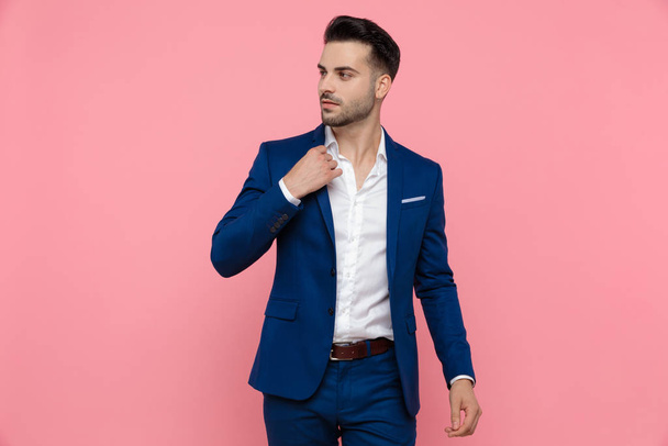 Charming businessman adjusting his collar and looking away while wearing blue suit and stepping on pink studio background - Zdjęcie, obraz