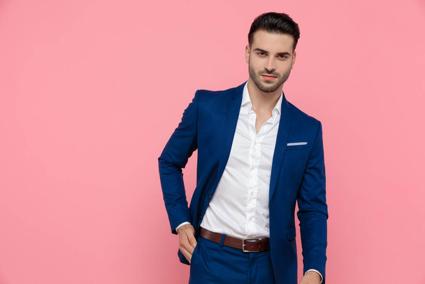 Jóképű üzletember fogja a kezét a zsebében, miközben kék öltönyt visel, és rózsaszín stúdió háttérben áll. - Fotó, kép