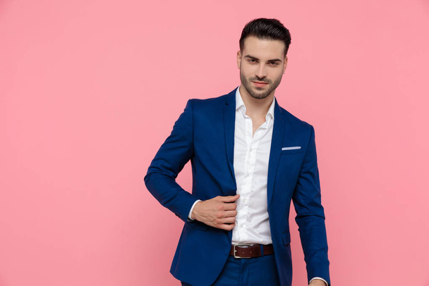 Γοητευτικός επιχειρηματίας χαμογελά και προσαρμόζει το σακάκι του φορώντας μπλε κοστούμι και στέκεται σε ροζ φόντο στούντιο - Φωτογραφία, εικόνα