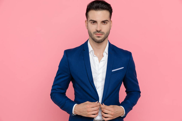 Confident businessman adjusting his jacket while wearing blue suit and standing on pink studio background - Foto, Imagem