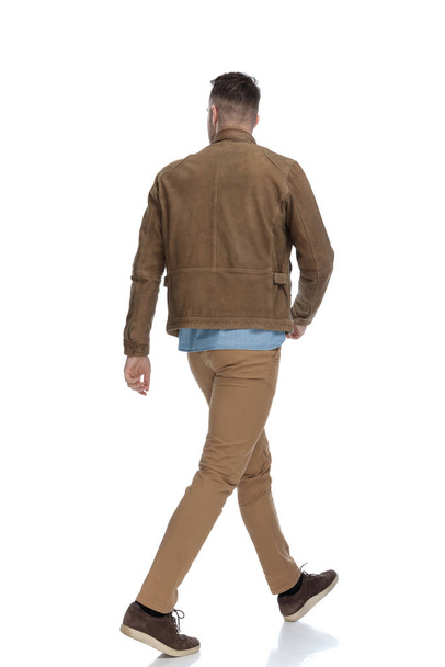 Вид ззаду жадібний випадковий чоловік у шкіряній куртці під час ходьби на білому фоні студії
 - Фото, зображення