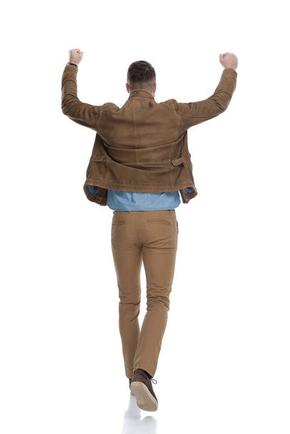 Vista trasera del hombre casual celebrando con la mano en el aire mientras usa una chaqueta de cuero y camina sobre fondo blanco del estudio
 - Foto, Imagen