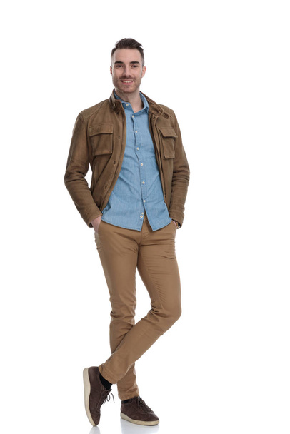 Positieve casual man glimlachen met beide handen in zijn zakken, terwijl het dragen van een leren jas en stappen op witte studio achtergrond - Foto, afbeelding